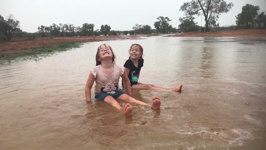 Two young girls sit, smiling, with wet hair, in an expanse of water on open country, as rain falls.