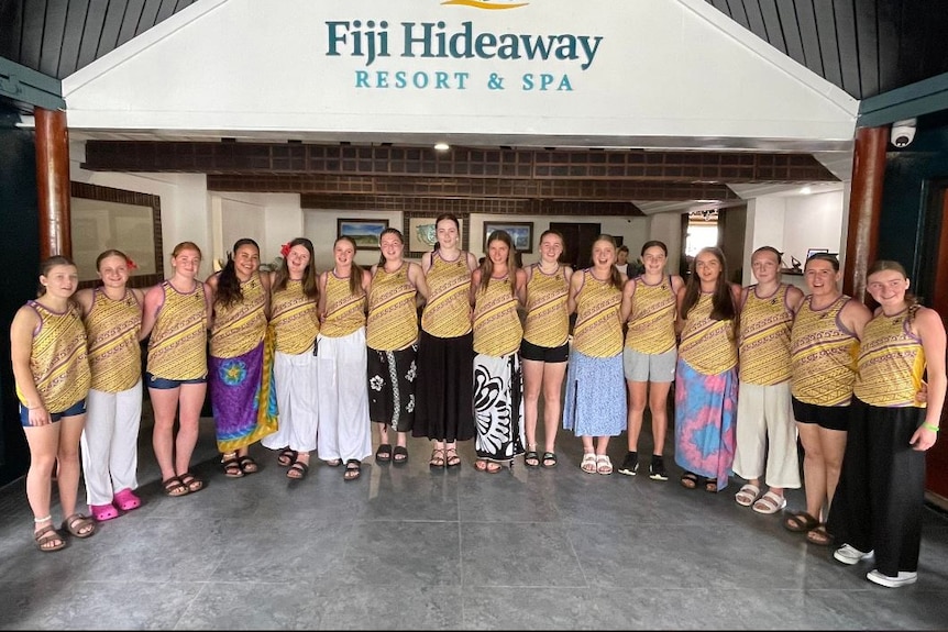 16 girls in matching shirts stand in front of a Fiji resort