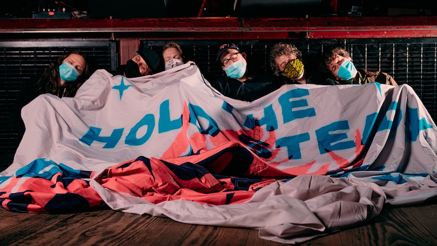 Six members of The Hold Steady sleep side by side up against a stage with a banner covering them. They each wear facemasks.
