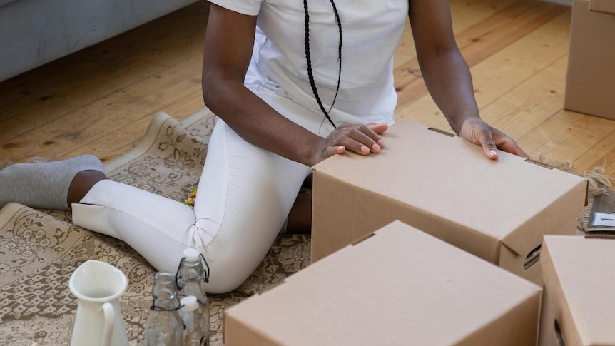 Woman packing boxes on living room floor, with glass bottles and a white vase beside one cardboard box.