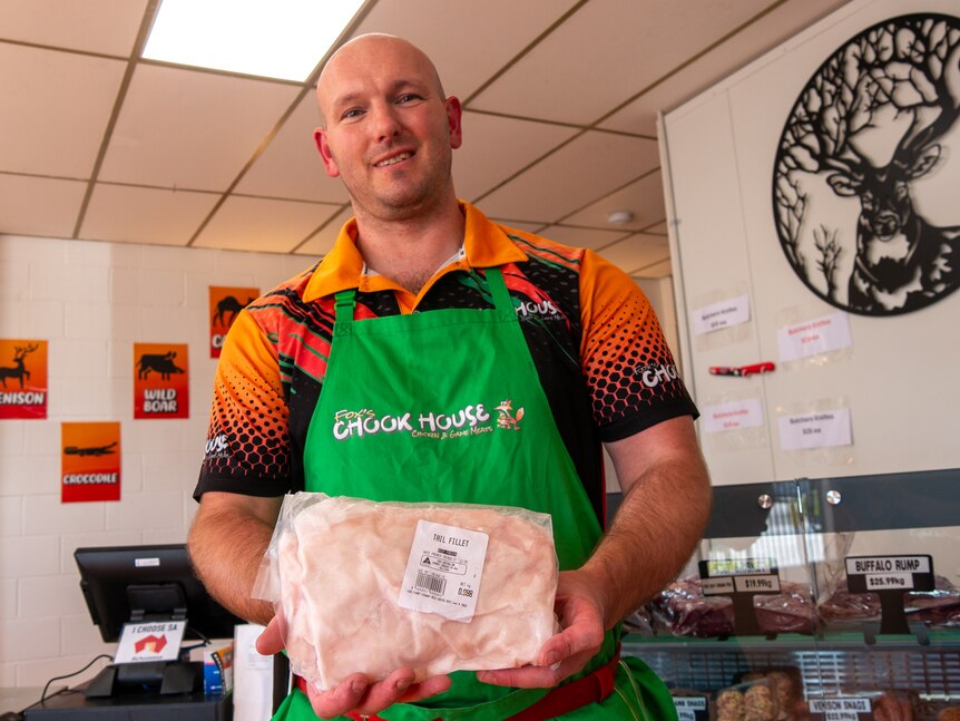 A smiling butcher stands in his shop, holding a package of meat.