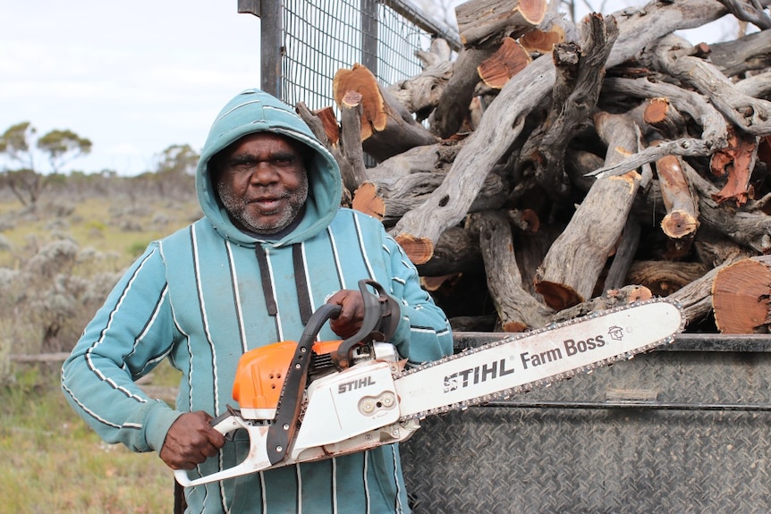 Aboriginal man in blue hoody, holding chainsaw in front of trailer of sawn wood.