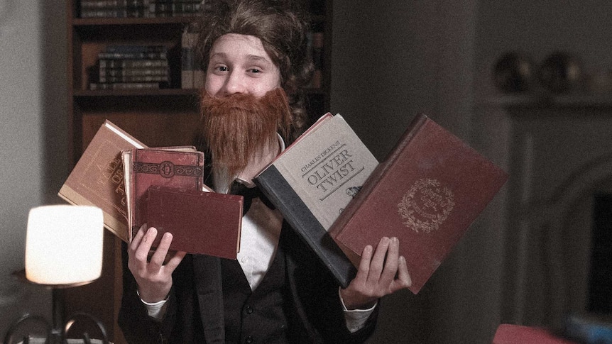A boy in vintage clothing holds up various books by Charles Dickens.