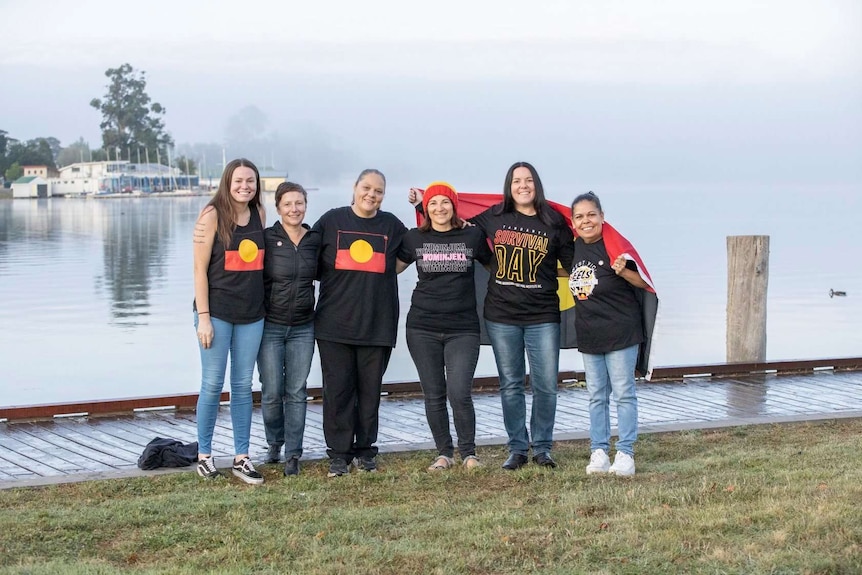 Six women, many wearing Aboriginal flags, stand together with Lake Wendouree behind.
