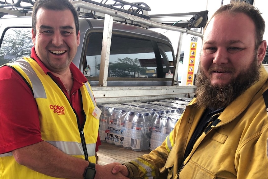 A Coles representative shakes hands with an RFS volunteer with water bottles on a ute in the background