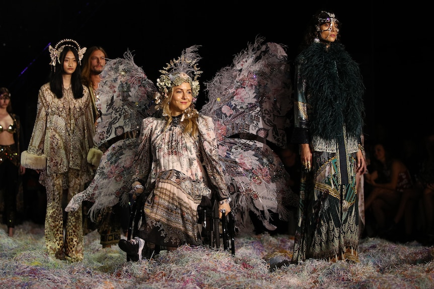 A model in a wheelchair with elaborate fair wings, other models stand around her