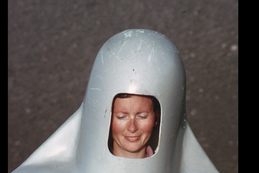 White woman with sun-burned cheeks smiles and looks downward putting on a giant silver helmet on a mountainside.