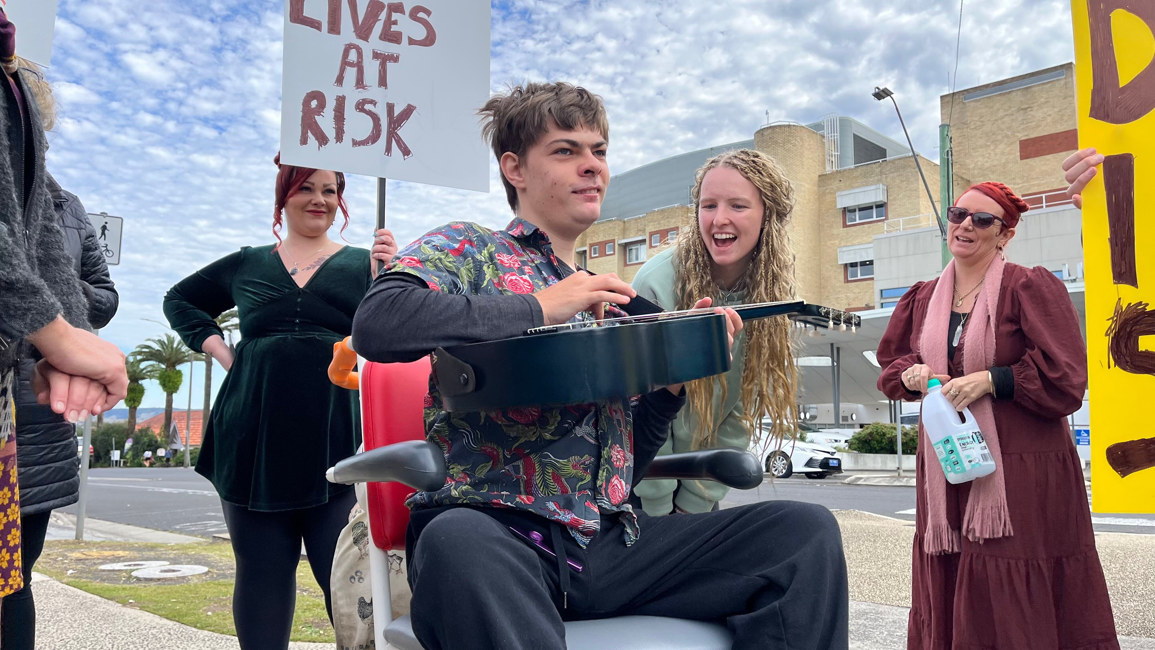 A young man sits in a wheelchair and holds a guitar while smiling, with a young woman next to him with a big grin on her face.