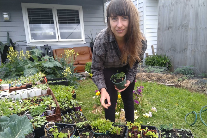 Young woman with long brown hair crouches down next to seedlings in her front garden.