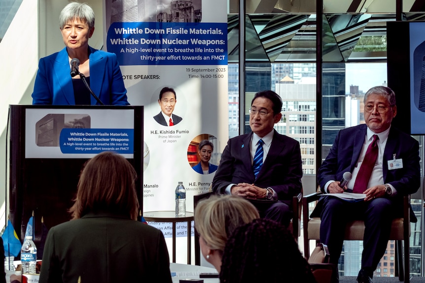 Penny Wong stands at a lectern on stage. Fumio Kishida and Enrique Manalo sit next to her.