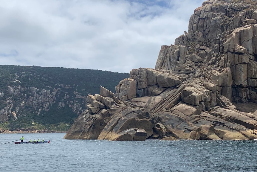 A surfboat rowing past a huge rock face.