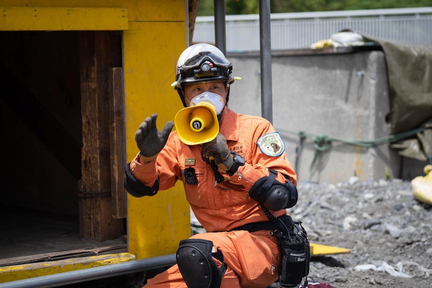 A man in orange jumpsuit and protective gear speaks into a yellow megaphone on an earthquake training site