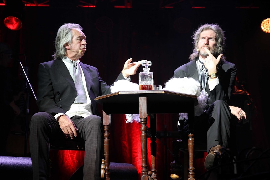 Two judges sit at a table with a decanter of alcohol in a stage production.