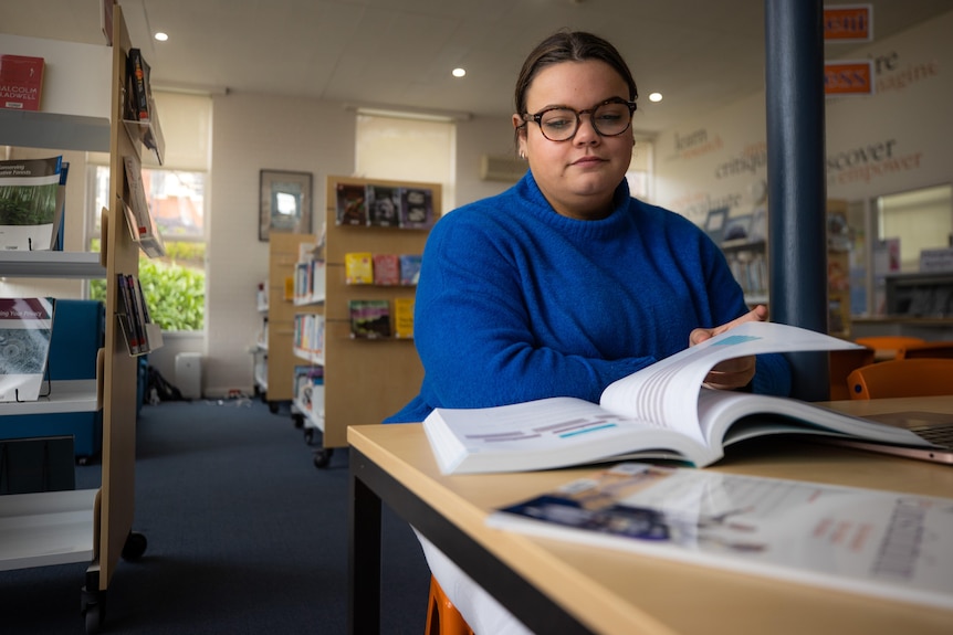 A young Indigenous woman in a blue jumper reads a book in a library