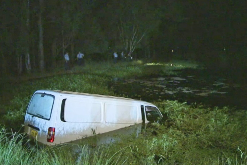 A van half submerged in a dam in the dark, with police in the background negotiating with a man in the water.