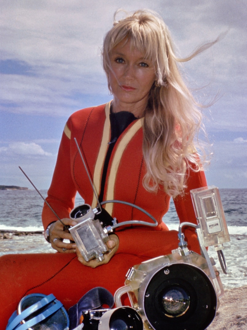 Archival photo of Valerie Taylor with long blonde hair wearing red wetsuit, posing with camera equipment.