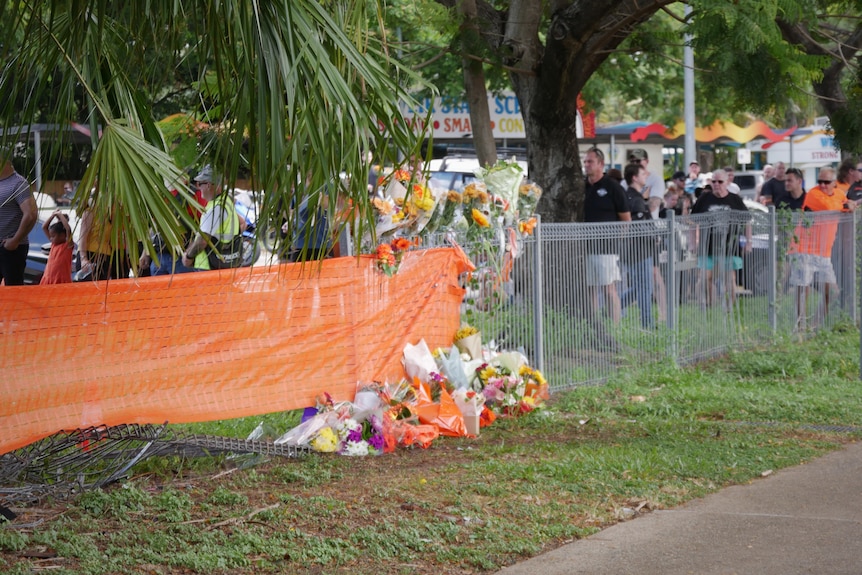 Orange flowers and tributes lay at a damaged metal fence. Behind the fence dozens of people are gathering.