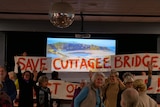 A bright red banner saying 'save cuttagee bridge' with people in front of it in a room.