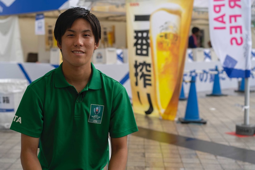 Shin Takuma smiles at the camera. In the background is a banner advertising beer.
