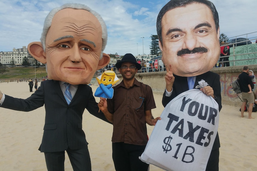 A man stands between people dressed as Malcolm Turnbull and Adani while holding an emoji sign. 