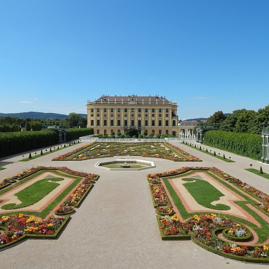 A well-manicured garden with intricate floral and hedge designs, with the facade of a large palace in the background.