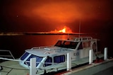 A boat moored by a small jetty with an orange fire burning in the background over the water.