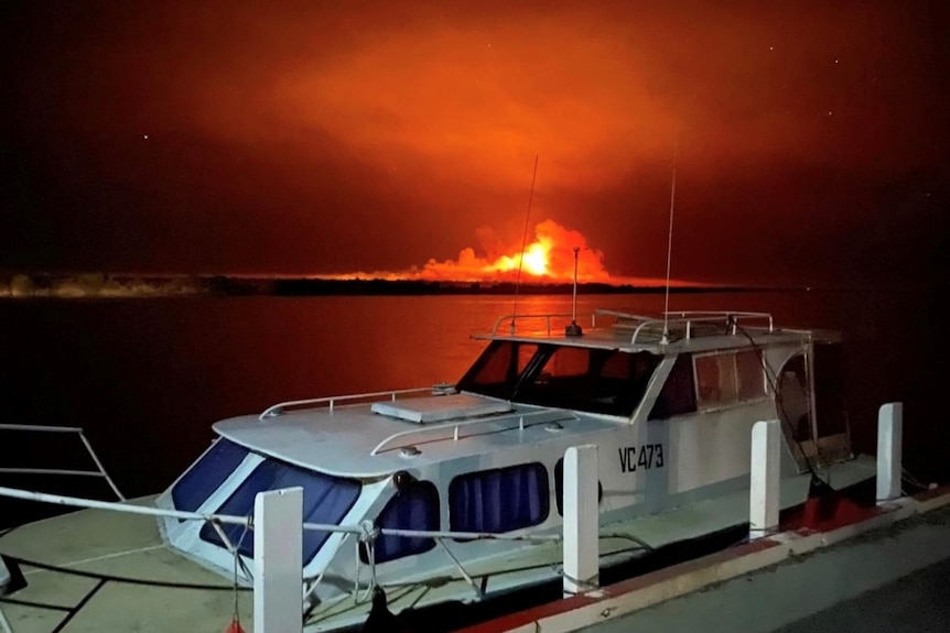 A boat moored by a small jetty with an orange fire burning in the background over the water.
