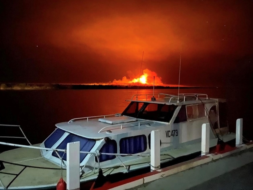 A boat moored by a small jetty with an orange fire burning in the background over the water.