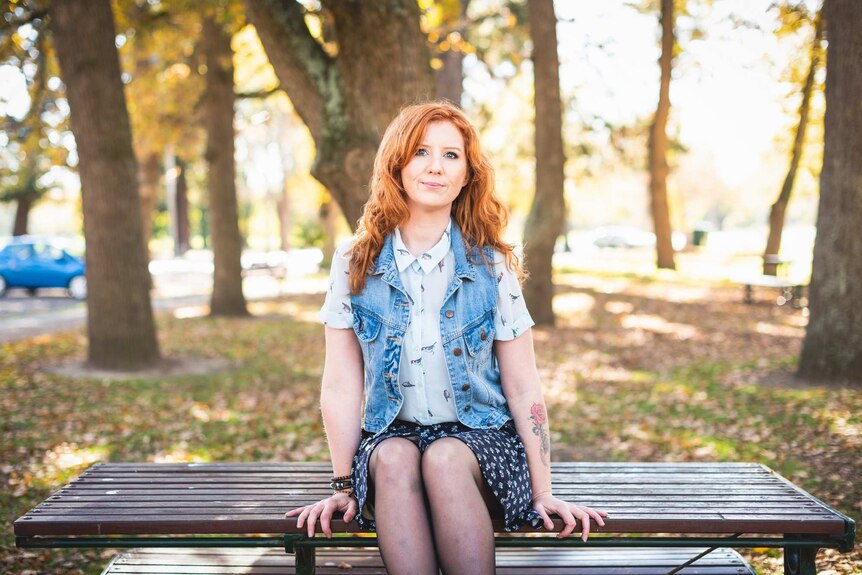 A woman with red hair sits on a bench with trees in the background.
