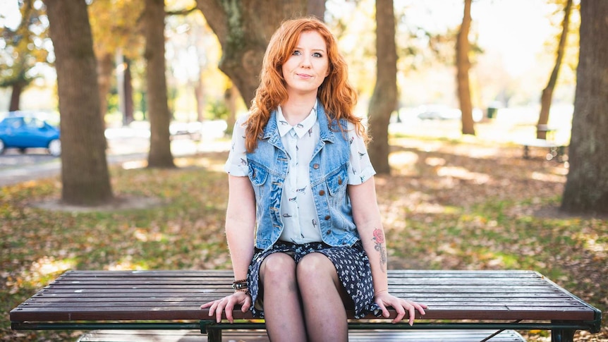 A woman with red hair sits on a bench with trees in the background.