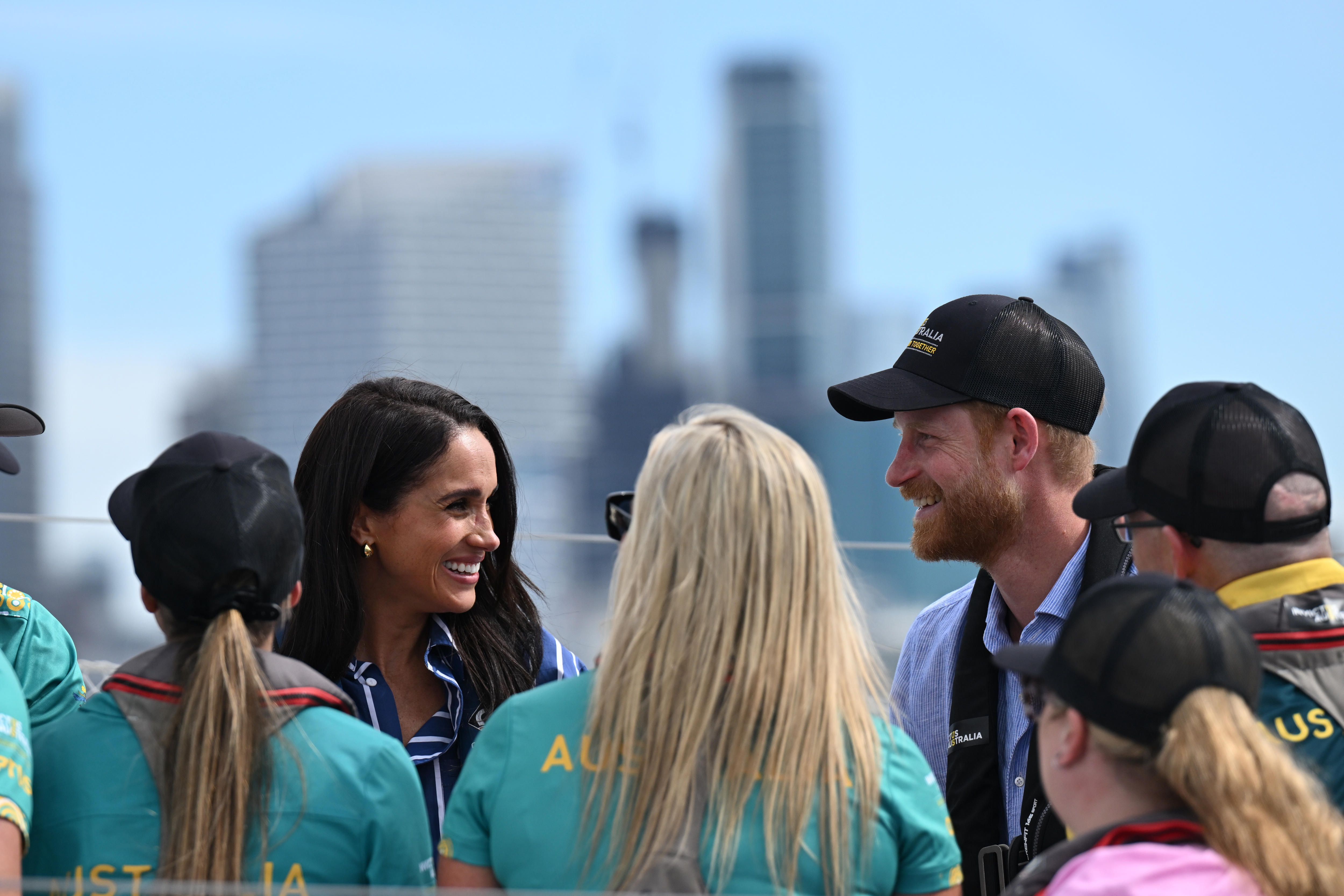 Harry and Meghan look at each other smiling in front of a group of people with the cityscape behind them.