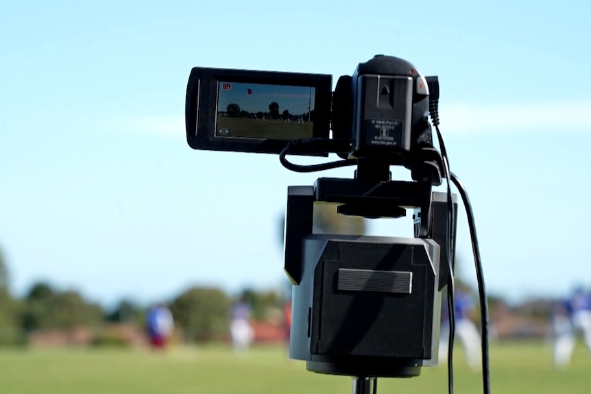 A small digital video camera sits on a stand outside. Out of focus in the distance is a cricket oval with players on it.