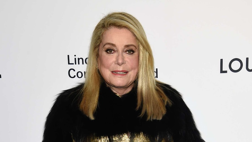 Medium shot of Catherine Deneuve standing in front of a publicity poster.