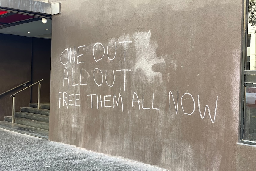 Chalk on the side of a building reads 'one out, all out, free them all'.