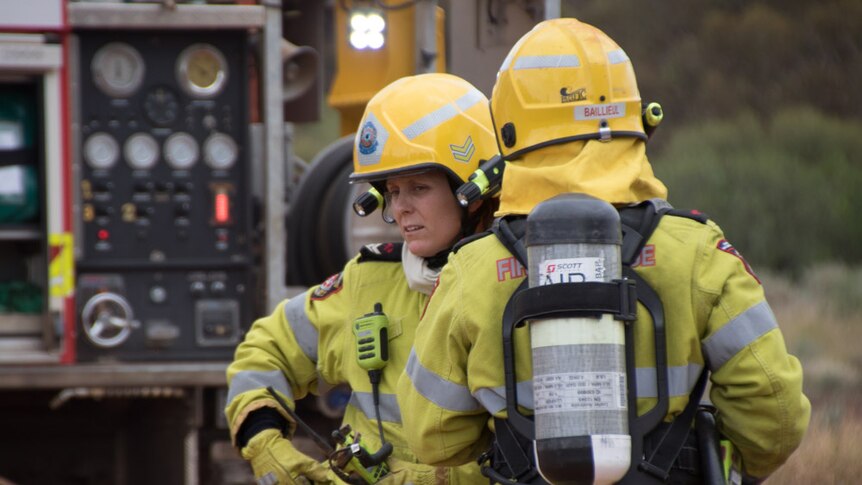 A woman in a high-vis fire fighting uniform complete with gloves and helmet chats to another firefighter behind a firetruck.