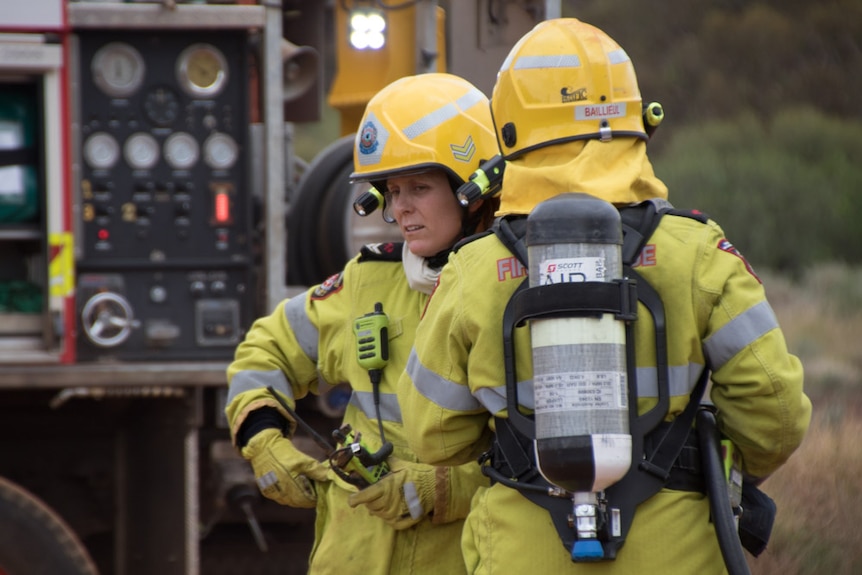 A woman in a high-vis fire fighting uniform complete with gloves and helmet chats to another firefighter behind a firetruck.