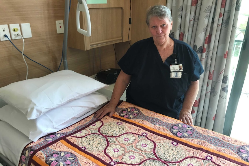 A woman stands beside a bed covered in a colourful patterned quilt in Palliative Care Ward in Wauchope.