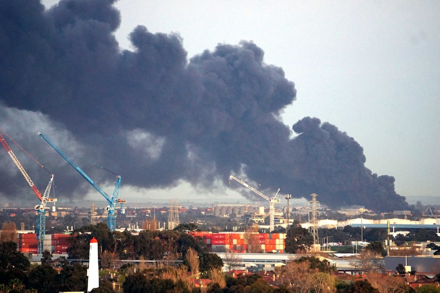 Thick black smoke fills the sky above cranes, shipping containers and industrial buildings.