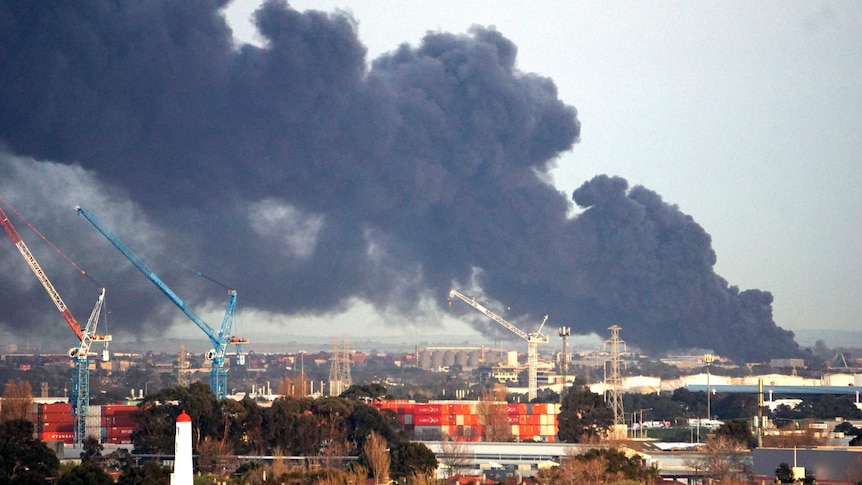 Thick black smoke fills the sky above cranes, shipping containers and industrial buildings.