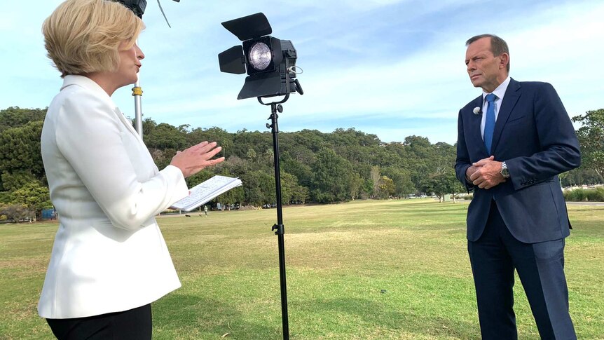 A woman wearing a white jacket and holding a notepad stands among lighting on a grassed area and speaks with a man in a suit.