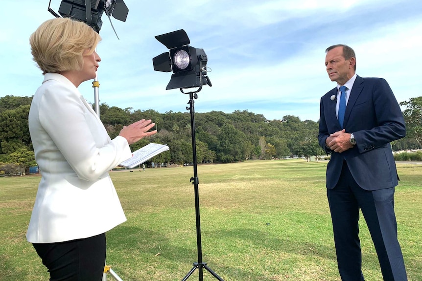 A woman wearing a white jacket and holding a notepad stands among lighting on a grassed area and speaks with a man in a suit.