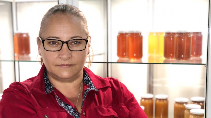 A woman with her arms crossed looks serious, with honey in jars on shelves behind her.