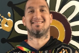 A bearded Steve Bastian stands in front of an Aboriginal artwork, smiling.