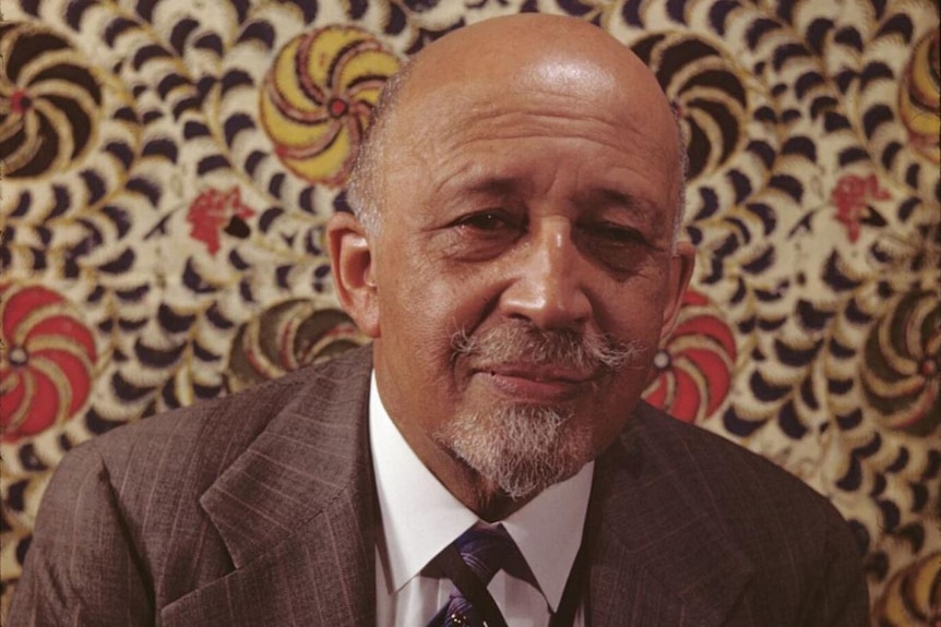 Against a patterned wall, a man in grey suit and tie, with twisted moustache and short beard, smiles subtly, looking ahead.