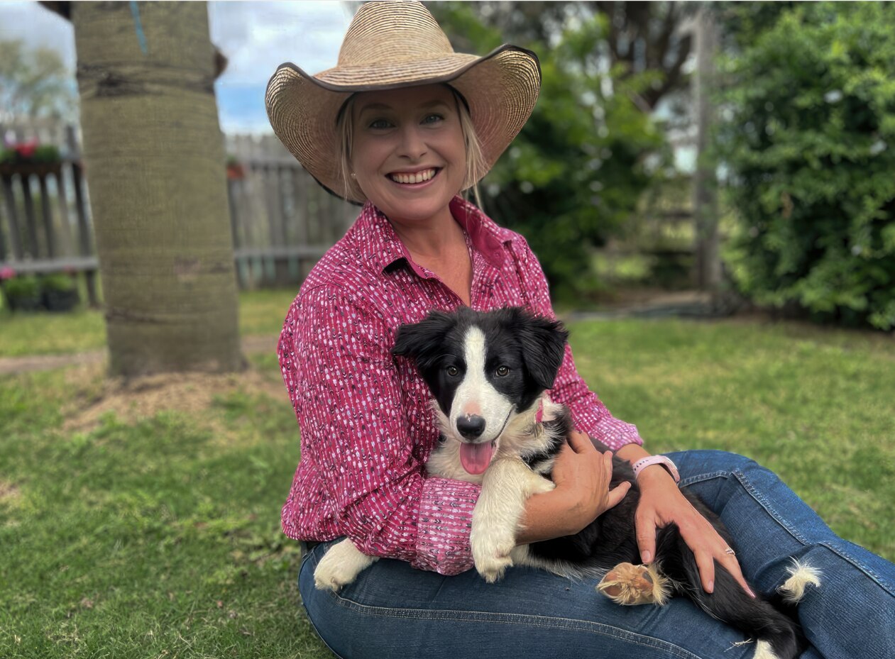 Woman in pink check shirt and broad-brimmed hat cuddles puppy and sits on the grass