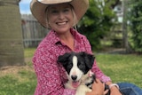 Woman in pink check shirt and broad-brimmed hat cuddles puppy and sits on the grass
