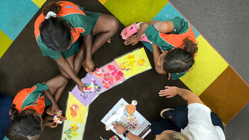 Three Indigenous children look at brightly coloured books with an adult.