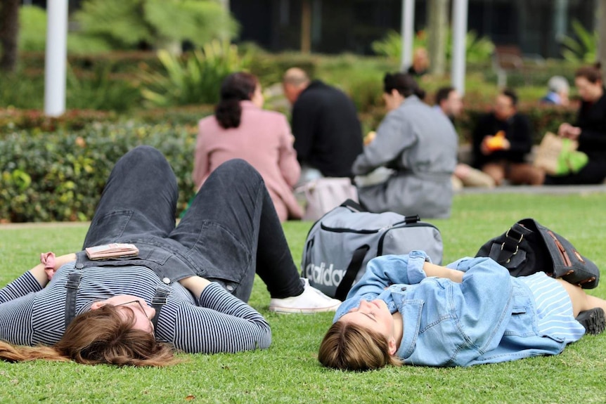 Two female friends lying down on grass together, with other people sat down eating their lunch in the background.