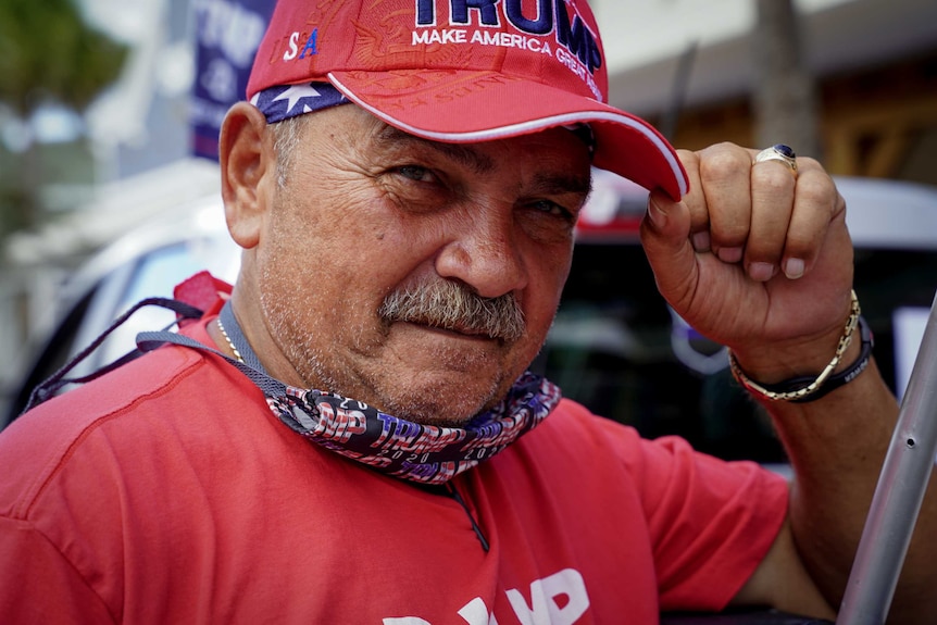 A man in a Trump cap, shirt and mask around his nick tweaks the brim