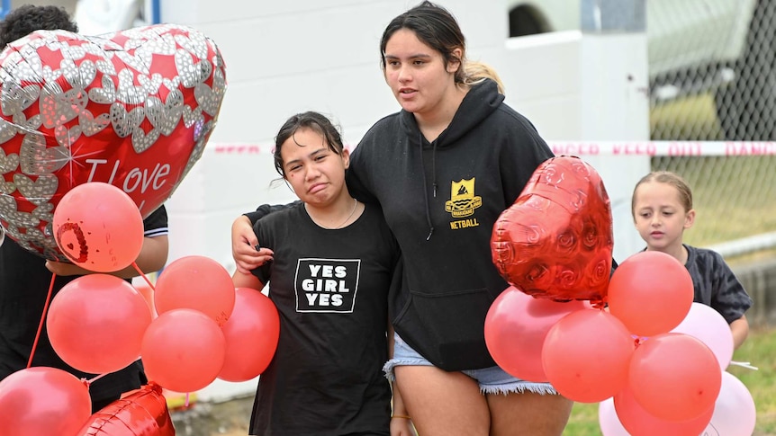 A teenager comforts a young girl crying as they bring red balloons to a blessing service